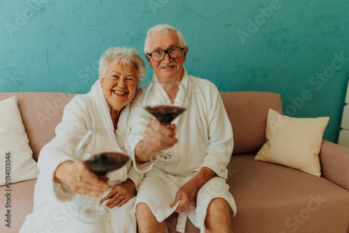 Tableau sur toile Happy senior couple sitting together in bathrobe on sofa with glass of wine