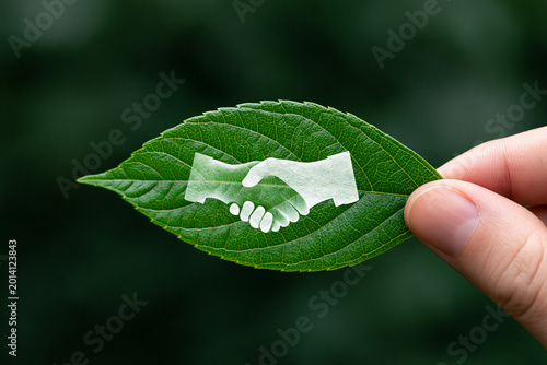 Conceptual image of environmental partnership showing a white handshake icon on a vibrant green leaf held by a hand against a blurred forest background representing sustainable business deals.