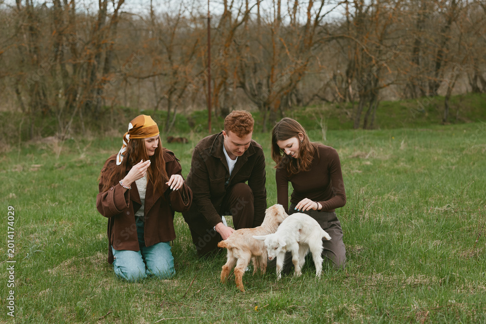 Naklejka premium Three young adults enjoy a spring day outdoors playing with two baby goats on green grass in casual warm clothing creating a relaxed and natural lifestyle moment.
