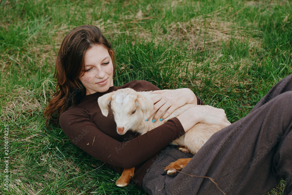 Fototapeta premium A goat and kid cuddle with a woman outdoors, a lifestyle moment of bonding and relaxation in a meadow. A tender, calm scene of companionship, nature, and simple joy in bright daylight.