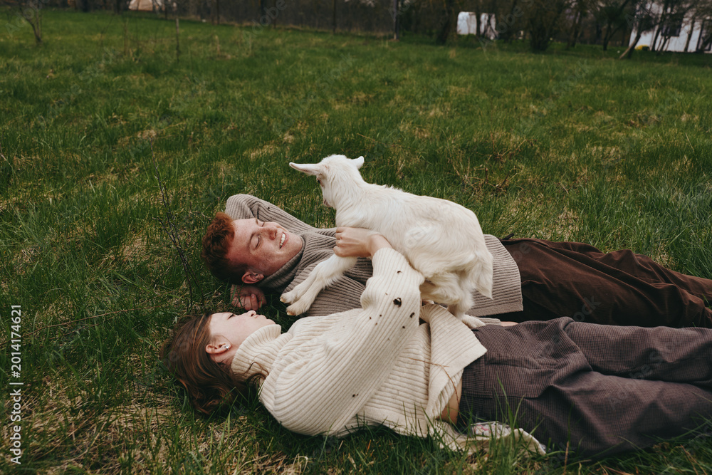 Naklejka premium Lifestyle moment with a couple lying on grass outdoors, a goat kid nearby, they smile and embrace, rustic calm scene, soft lighting, candid everyday joy, harmony between humans and animals.