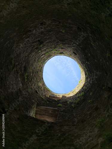 Looking up from inside a rough stone well or shaft, the circular opening frames a blue sky while small plants grow along the weathered walls.