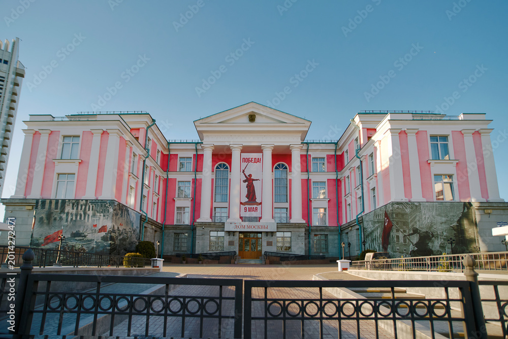 Naklejka premium Minsk, Belarus. May 5, 2026. Dom Moskvy cultural center adorned with large scale war memorial paintings, Victory Day celebration banner between white columns during bright spring day, House of Moscow