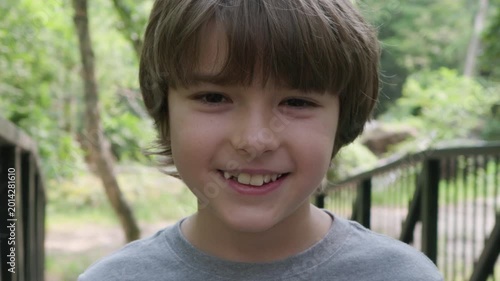 Portrait Young Boy Smiles Outdoors on Bridge in Lush Green Park. Pleasure Relax of Walking in Forest. Happy and Tranquility Little Child Looking at Camera in Nature. Children Healthy Lifestyle.