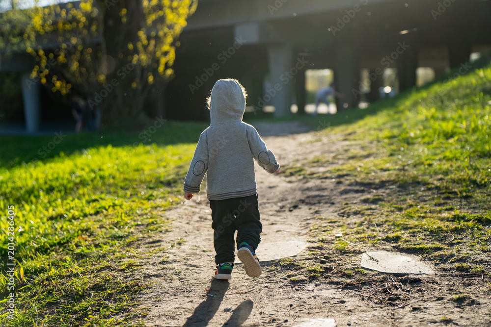 Fototapeta premium Small boy in hoodie walking on a sandy path in a city park environment featuring green slopes and concrete pillars representing childhood curiosity and urban outdoor play