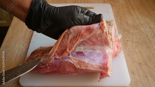 Fotografie Hands wearing black protective gloves use a sharp knife to carefully slice and trim a large piece of raw red meat on a white plastic cutting board during a professional butchering process