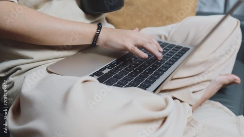 Person sitting casually on a couch using a laptop for remote work or e learning, typing and browsing from home to enjoy the comfort and flexibility of hybrid lifestyle