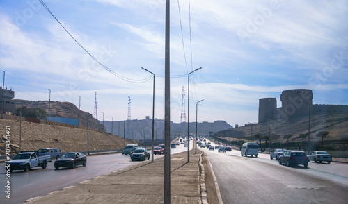 A view from a tourist bus showing heavy traffic on a highway leading towards the historic Khan el-Khalili market in Cairo, Egypt, with ancient stone structures visible in the background.