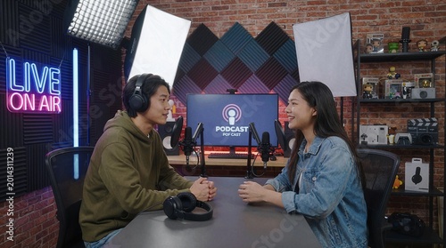 Two people recording a podcast at a desk in a studio with a Live On Air neon sign