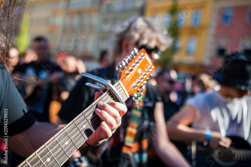 Close-up shot of a musician playing a 12-string acoustic guitar with a capo during an outdoor street performance, focusing on the left hand and the headstock.