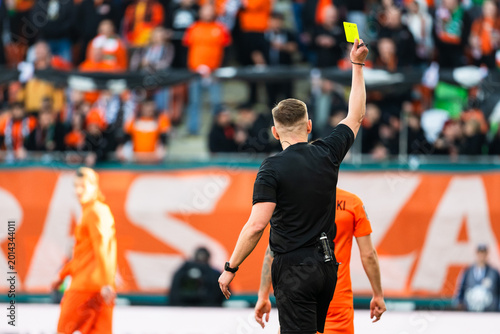 Back view of a soccer referee in a black uniform holding up a yellow card for a foul during a professional match in a crowded stadium.