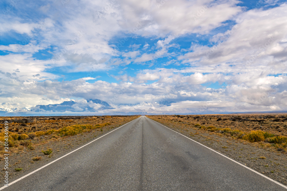 Fototapeta premium Straight asphalt road leading towards distant mountains under a blue sky with dramatic clouds in Patagonia landscape