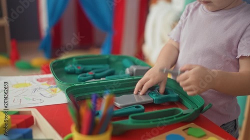 Girl playing with green toy medical kit, holding plastic pliers with bare hands in preschool classroom; playful curiosity.