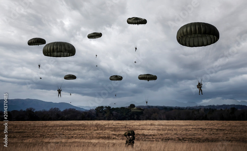 Military paratroopers in green parachutes descend toward a vast dry field under a moody, overcast sky, representing strategic deployment, tactical training, and airborne operations.