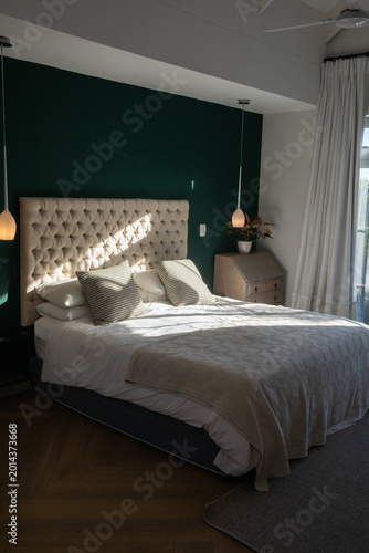 Double bed with tufted beige headboard sitting in center of bedroom, catching sun through curtains