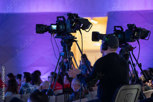 Professional camera operator filming corporate conference, live video production with crowd, tripod, headset, purple blue lighting.