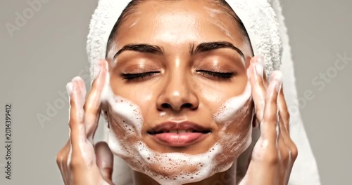 Woman applying white foam facial mask with towel wrap for spa skincare and relaxation beauty treatment