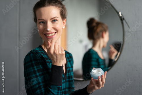 Young woman smiling at the camera while applying facial moisturizer cream to her face, having a playful dot on her nose, standing in a modern bathroom with a mirror