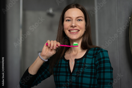 Young woman smiling while holding a toothbrush with toothpaste, preparing for her daily oral hygiene routine in the bathroom, emphasizing dental care and a fresh start