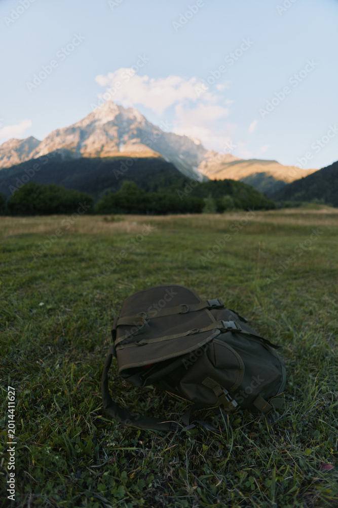Fototapeta premium A lone backpack sits on a grassy meadow with towering mountains in the background, capturing a moment of outdoor exploration and quiet solitude.