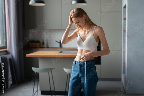 Young woman in a modern kitchen looks distressed, holding her head while measuring her waist with a tape, reflecting anxiety, body image struggle and dieting pressure