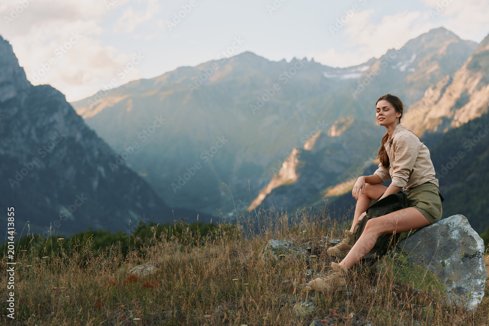 Naklejka premium Woman sits on a rock in a mountain meadow, enjoying the view. Outdoor lifestyle scene highlighting nature, solitude, and calm.