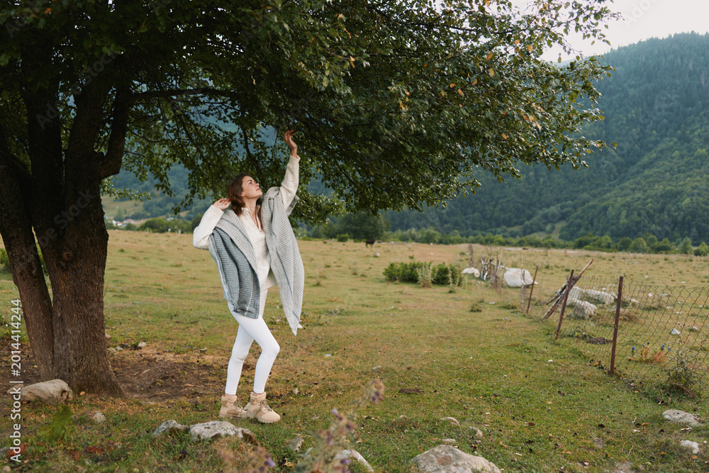 Fototapeta premium A woman in a light cardigan dances beside a tree in a rural field, capturing a carefree outdoor moment and a sense of freedom, a lifestyle scene of wellness and nature.
