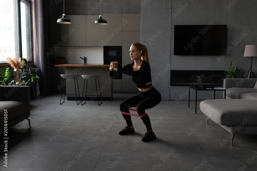 Fototapeta premium Young woman is performing a squat exercise with a fitness resistance band in the living room of her modern apartment, focusing on health, wellness, and a fit lifestyle