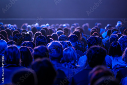 Large seated audience bathed in blue stage lighting, focused on live performance, shared experience, and collective anticipation.