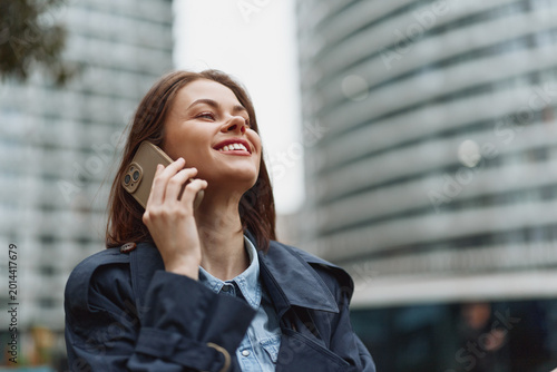 Φωτογραφία Young woman in a navy coat chats on a smartphone in an urban plaza, smiling as tall buildings rise behind, capturing a candid city moment and lively energy