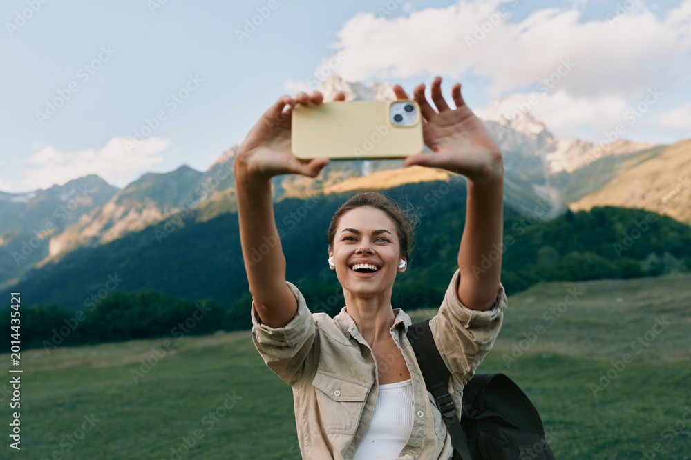 Naklejka premium Young woman lifestyle traveler takes selfie with smartphone on mountain meadow background, smiling happy tourist enjoys nature and outdoor adventure in sunny day.