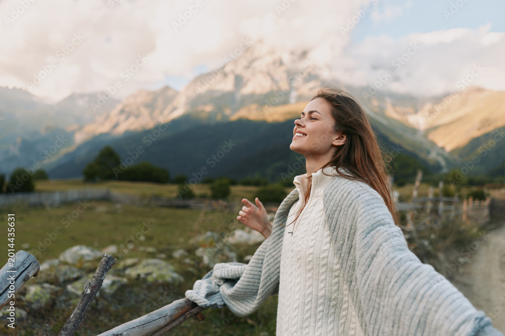 Fototapeta premium A woman stands outdoors on a sunny mountainside, arms outstretched, smiling, wearing a cozy sweater. Bright landscape highlights freedom, happiness, and a carefree lifestyle.
