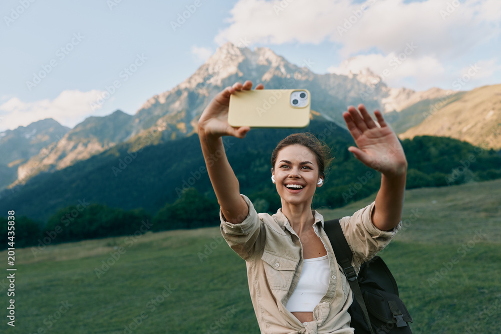 Naklejka premium Cheerful woman outdoors taking a selfie in a mountain meadow, bright smile, casual outfit, sunny day, sense of adventure, natural colors, scenic backdrop amid lush greens