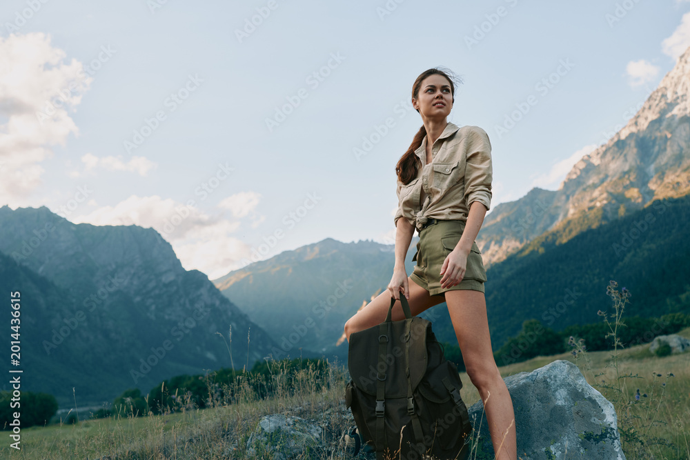 Fototapeta premium Woman outdoors hiking with backpack beside a mountain landscape in sunlit scenery, casual outfit, confident pose, natural light, serene meadow background