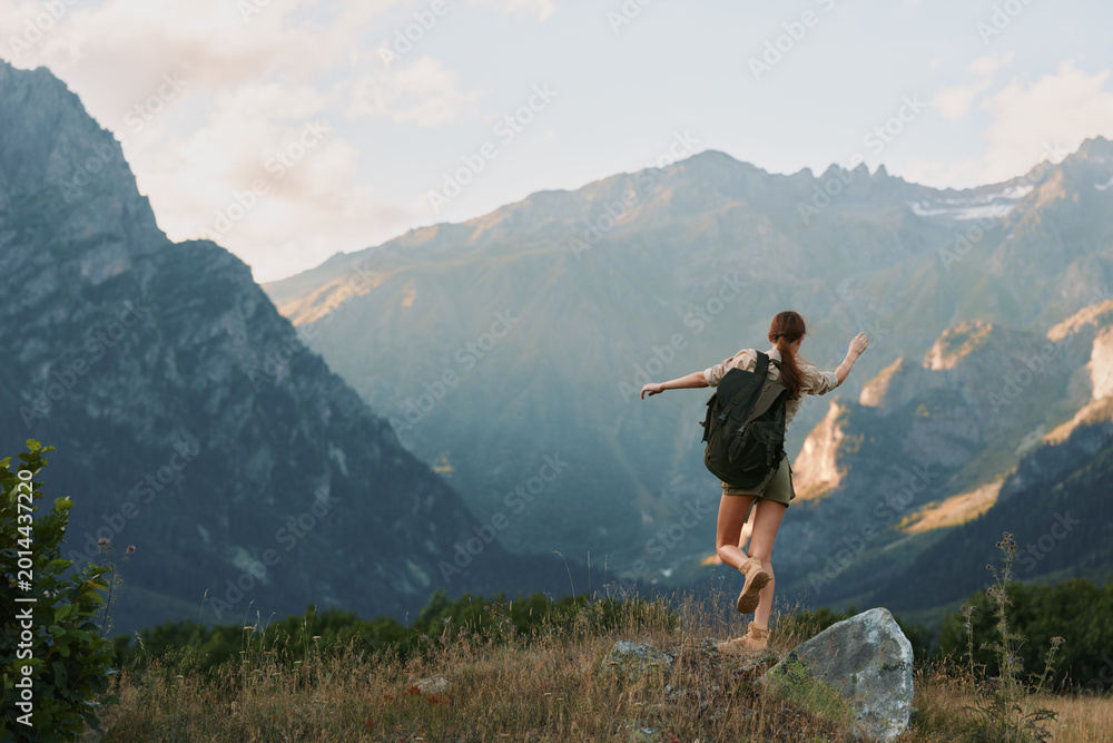 Fototapeta premium A young woman with a backpack strides through a grassy meadow toward dramatic mountains, radiating adventure, freedom, and focus on the trail ahead.