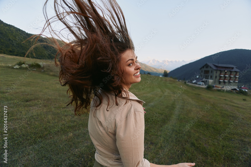 Naklejka premium Happy woman outdoors turning with hair flying, smiling joyfully, wearing casual clothes, green meadow, mountain landscape, clear sky background
