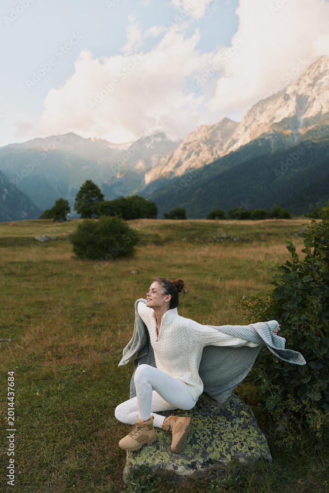 Fototapeta premium A woman sits on a rock in a white knit sweater and boots, stretching her arms, gazing toward distant mountains, a calm outdoor lifestyle moment in nature.