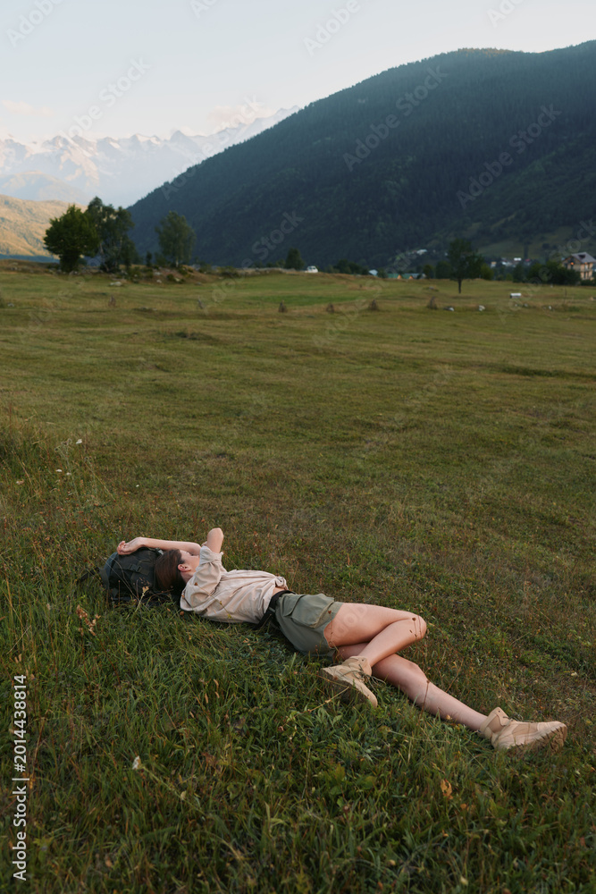 Naklejka premium Lying woman in a sunlit meadow, relaxed pose in a wide field with distant mountains, celebrating outdoor lifestyle, natural colors and peaceful countryside under blue sky warm breeze vivid greens.