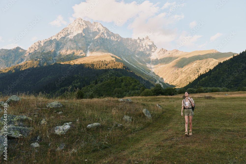 Fototapeta premium Woman hiker in a mountain meadow, exploring rugged terrain with a backpack, shorts and boots, gazing at distant peaks, a calm outdoor scene with solo travel vibe.