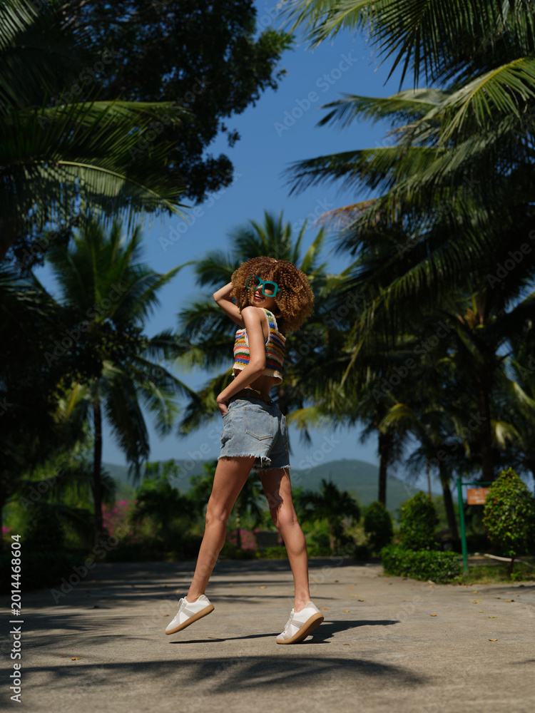 Naklejka premium young woman with curly hair wearing sunglasses and denim shorts jumping on a sunny tropical path surrounded by palm trees and clear blue sky