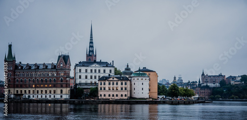 Stockholm Sweden Riddarholmen Waterfront Skyline with Historic Buildings and Church Spire Reflected in Calm Water Under Overcast Sky