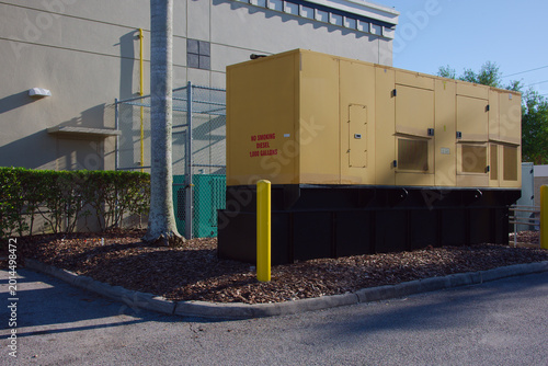 Large yellow diesel backup generator sits on a raised platform beside a commercial building, surrounded by mulch, safety bollards and fencing, illustrating infrastructure, emergency power and industri