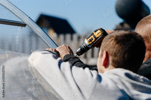 two men workers screw transparent polycarbonate sheet to metal frame with screwdriver, close-up view of hands with tool, installing greenhouse, seasonal gardening, sunny summer day