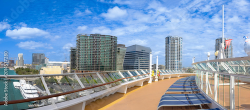 Panoramic view from a cruise ship deck in Tampa, Florida, USA. Sun loungers, glass railing, and modern downtown skyscrapers under a bright blue sky with clouds.
