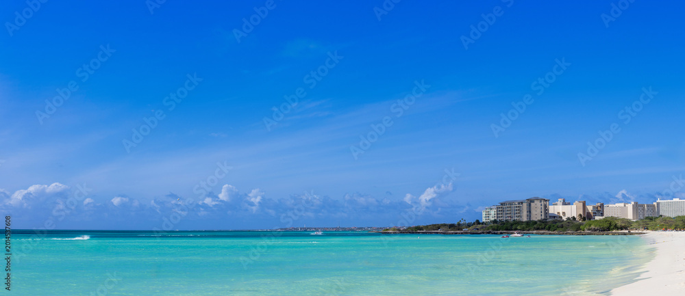 Fototapeta premium Panoramic view of Eagle Beach in Aruba featuring turquoise Caribbean water and a white sand coastline with a tropical resort skyline under a clear blue sky