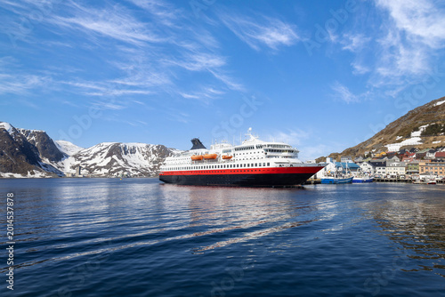 A large Norwegian Cruise ship with red hull docked at Honningsvag port, Norway, with snow-covered mountains and a small Arctic town visible under a bright blue sky