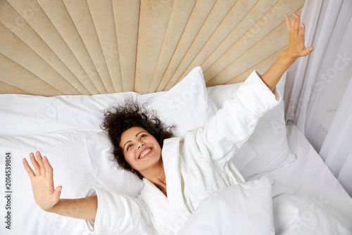 Φωτογραφία Smiling Woman Waking Up In Bed And Stretching Her Arms Up