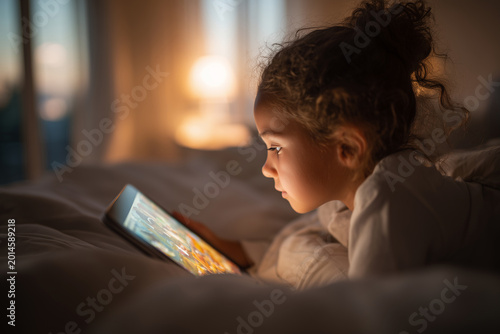 Young girl lying in bed and using a digital tablet in warm evening light