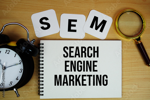 SEM Search Engine Marketing text written on notepad with magnifying glass and alarm clock on wooden background