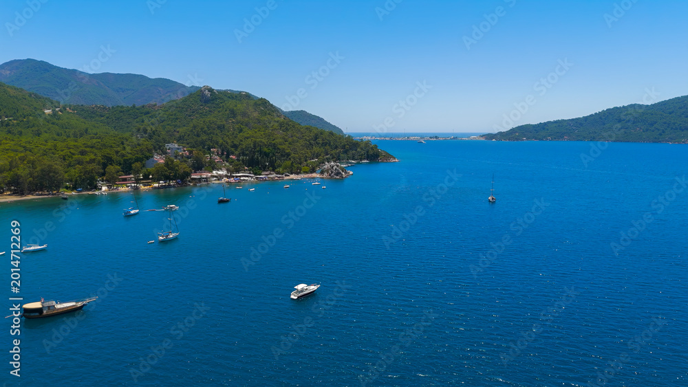 Fototapeta premium Marmaris, Turkey. Aerial drone flight along lush green forest coastline in Marmaris Bay. Sandy beaches, yachts sailing on blue water and coastal road on a sunny day.. Aerial View